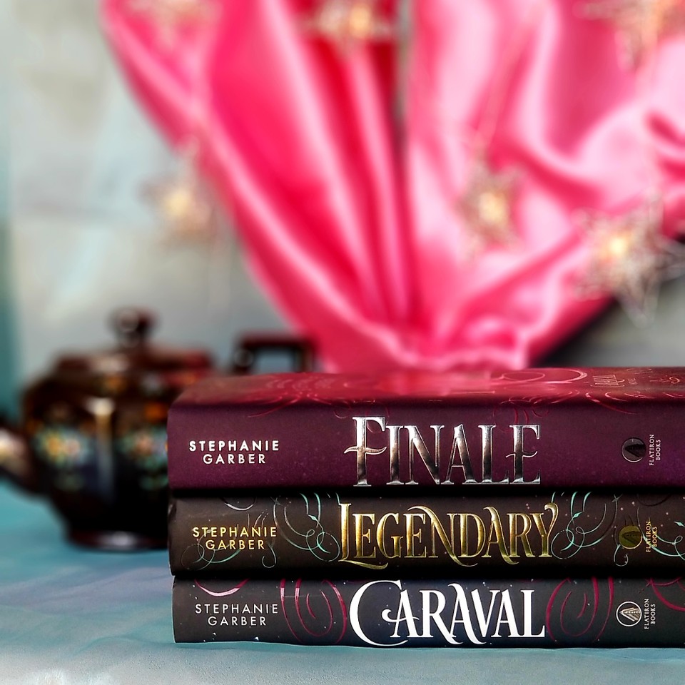 Stack of all three Caraval young adult trilogy books with pink and light blue fabric in the background and an antique teapot