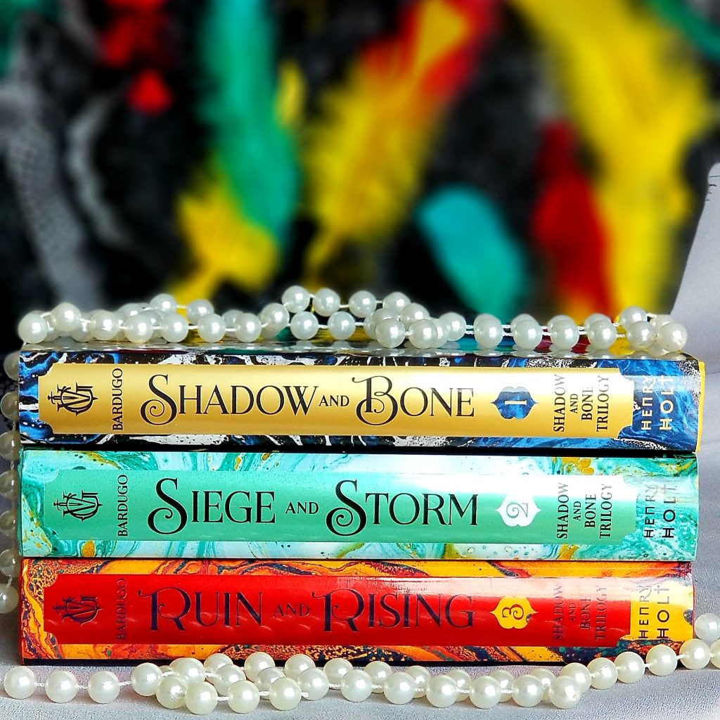 Stack of all three Shadow and Bone young adult trilogy books with multi-colored feather background and pearls